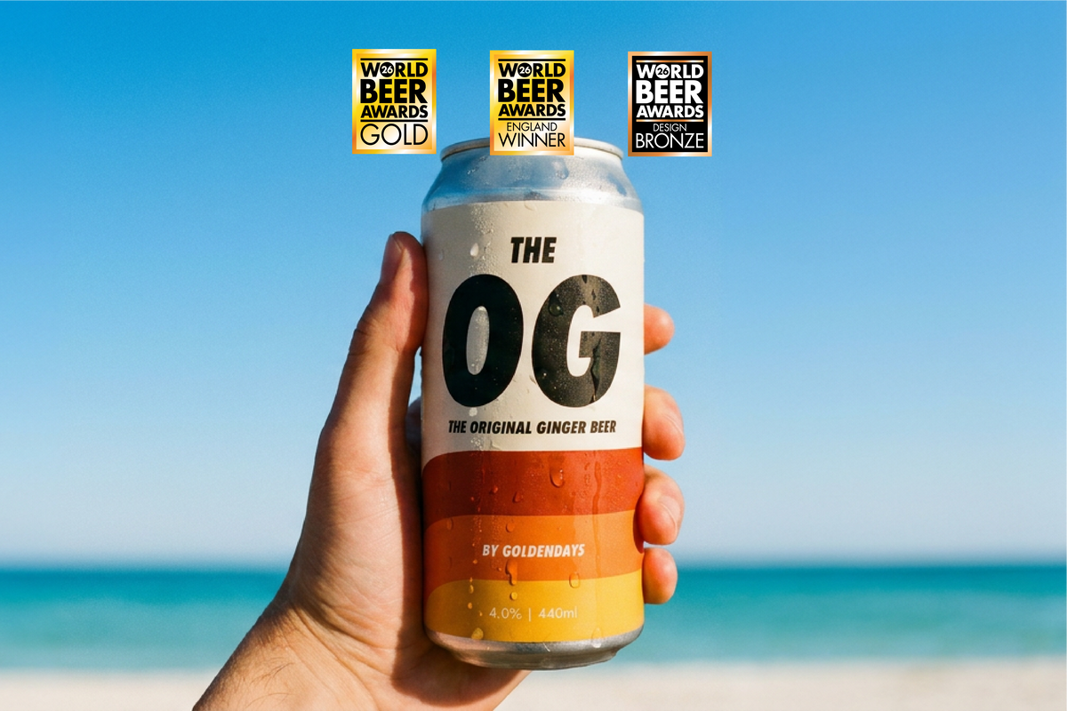 A hand holds a can of OG Gold Blonde beer in front of a sunny beach background with blue sky and ocean. Three award badges for 'World Beer Awards' are displayed at the top of the can.